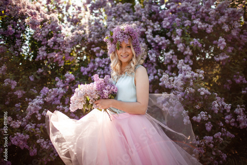 Portrait of a beautiful girl with a wreath on her head made of a purple flowers. A woman dressed pink skirt and blue sleeveless top walks outside against the background of flowering purple trees