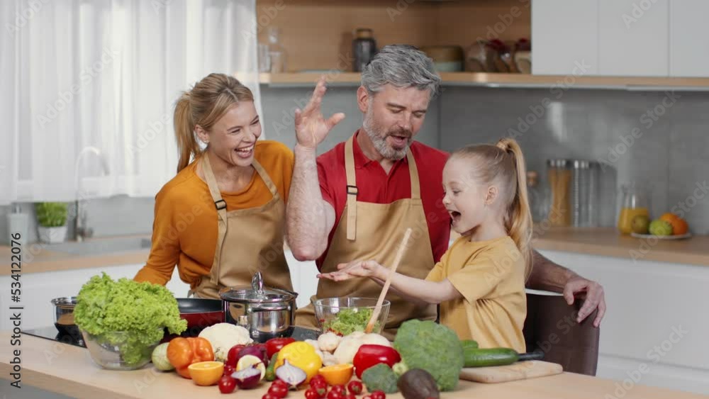 Fun at kitchen. Happy family of three preparing dinner together at kitchen, laughing and mixing salad, tracking shot Fun at kitchen. Happy family of three preparing dinner together at kitchen, laughing and mixing salad, tracking shot