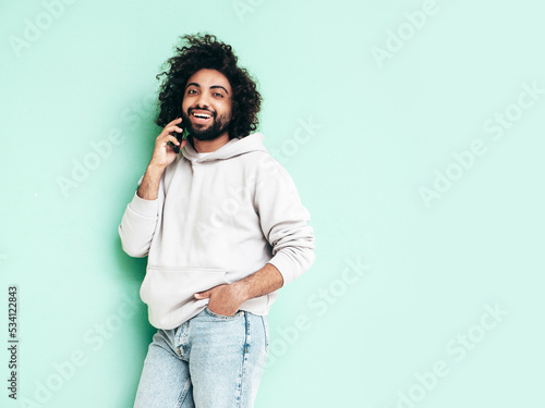 Handsome smiling hipster  model.Unshaven Arabian man holding smartphone. Fashion male with long curly hairstyle posing near green wall in studio. Talking at call