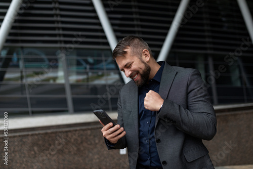 businessman in a classic suit on the background of an office building with a mobile phone in his hands energetic and emotional clenching his hand into a fist looking at the phone screen with a smile