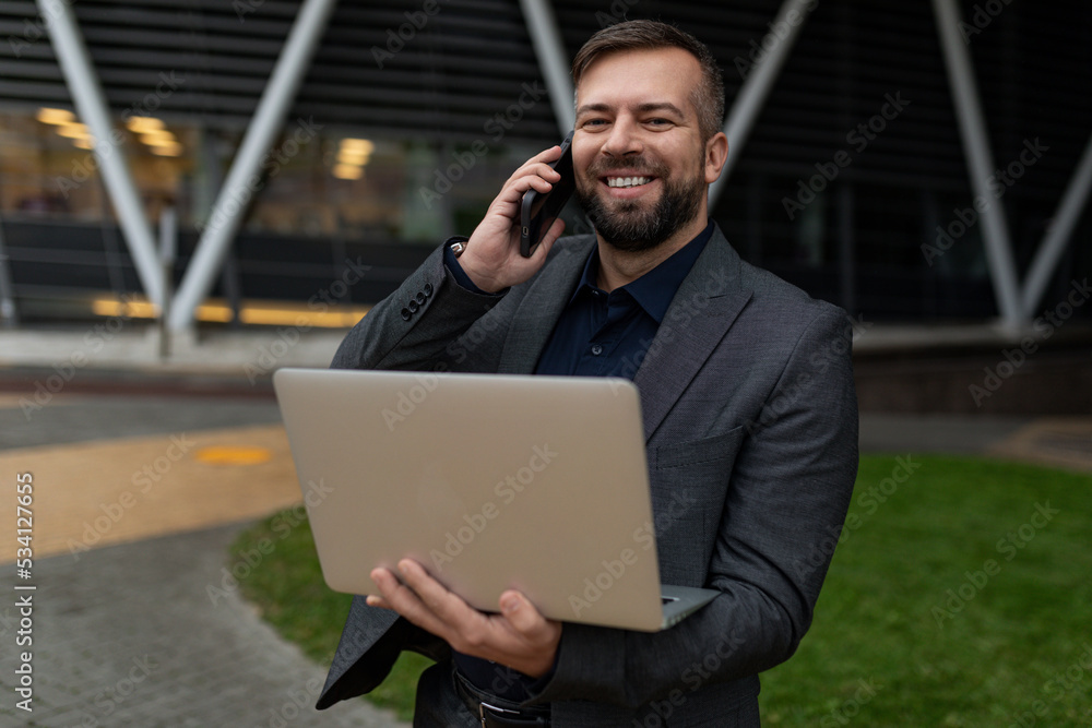 pretty adult man in a business suit with a laptop in his hands speaks on a mobile phone on the background of the office center, Concept of a successful realtor pretty adult man in a business suit with a laptop in his hands speaks on a mobile phone on the background of the office center, Concept of a successful realtor
