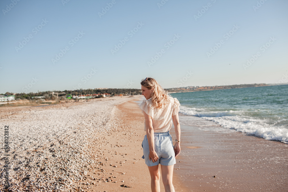 Back view of  beautiful girl dressed in a blue shorts and a white light blouse runs on a deserted beach, enjoys freedom and loneliness and look at the biew of the ocean