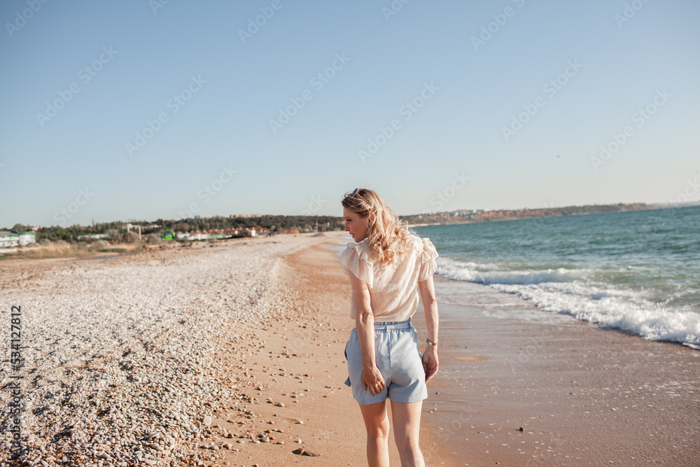 Back view of beautiful girl dressed in a blue shorts and a white light blouse runs on a deserted beach, enjoys freedom and loneliness and look at the biew of the ocean Back view of beautiful girl dressed in a blue shorts and a white light blouse runs on a deserted beach, enjoys freedom and loneliness and look at the biew of the ocean