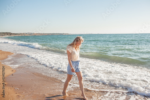 A beautiful girl dressed in a blue shorts and a white light blouse runs on a deserted beach, enjoys freedom and loneliness and look at the biew of the ocean photo