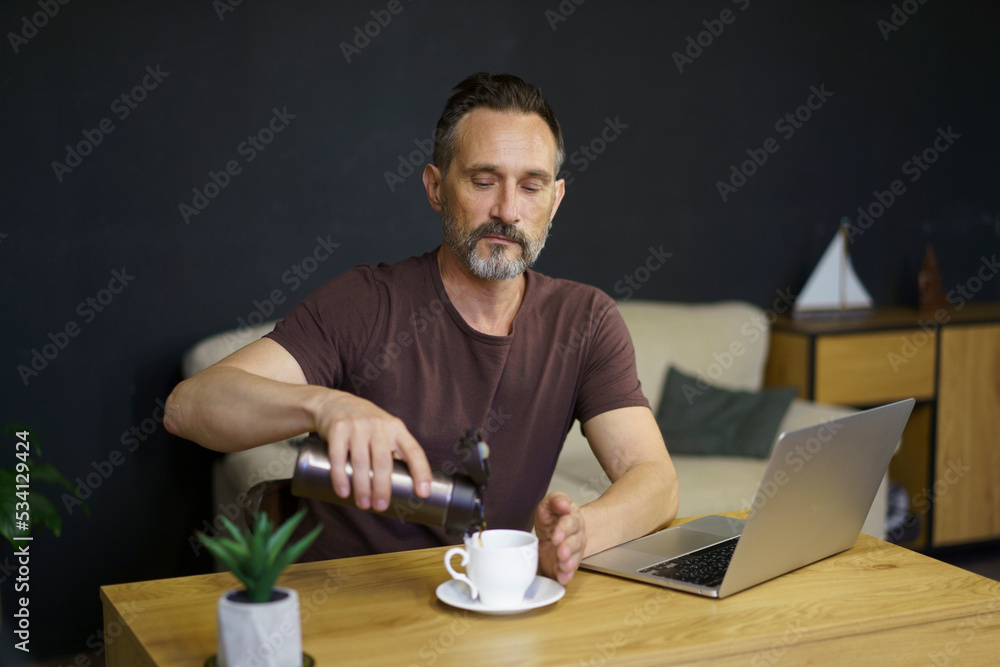 Bearded middle-age man pouring coffee from thermos to cup sitting at home working place with laptop Bearded middle-age man pouring coffee from thermos to cup sitting at home working place with laptop