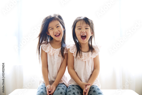 2 cute little lovely asian sisters sitting at home together and laughing to camera