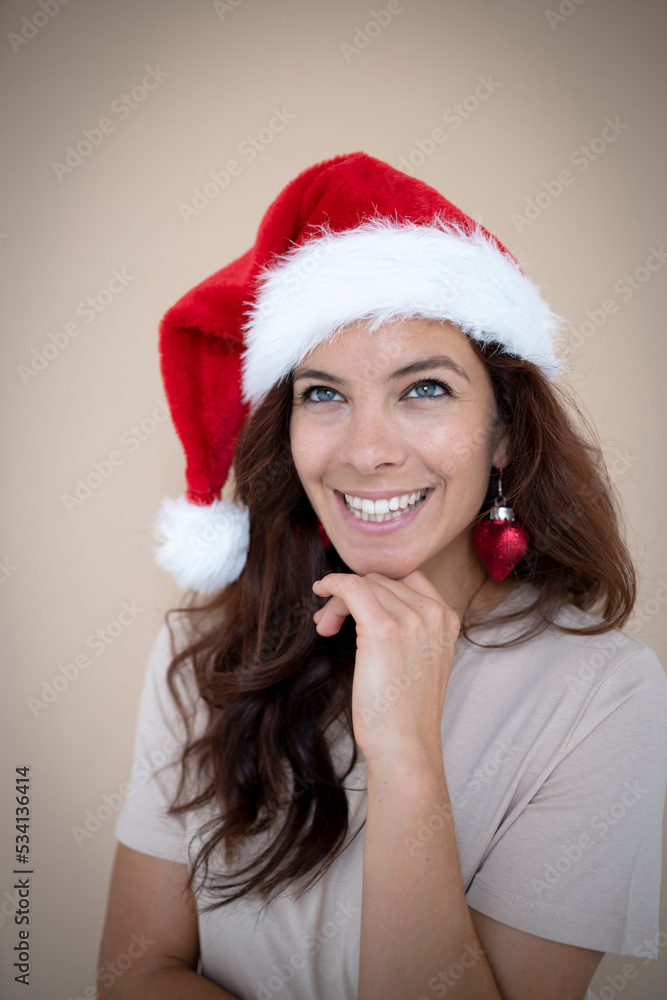 Pretty young woman with dark hair wearing Christmas hat and heart earrings in the shape of Christmas tree ball in front of brown background Pretty young woman with dark hair wearing Christmas hat and heart earrings in the shape of Christmas tree ball in front of brown background