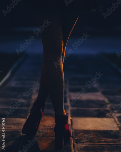 Women's legs in fishnet stockings close-up on a night street, in the light of street lamps. Back view.