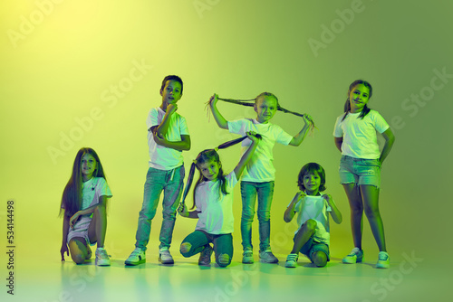 Group of children, little girls and boys in sportive casual style clothes dancing isolated on green background in yellow neon light.