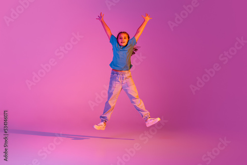 Expressive little girl, kid in casual bright clothes jumping isolated over pink background in neon. Action, dance, happy childhood