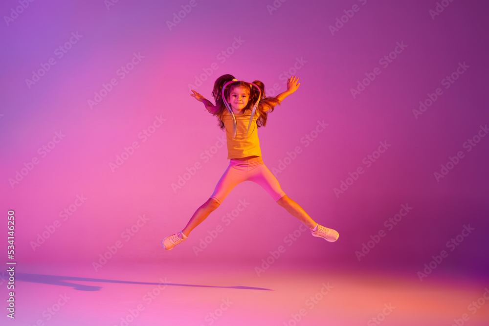 Expressive little girl, kid in casual bright clothes jumping isolated over pink background in neon. Action, dance, happy childhood Expressive little girl, kid in casual bright clothes jumping isolated over pink background in neon. Action, dance, happy childhood