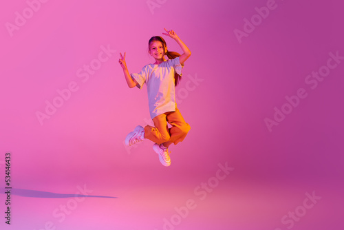 Beautiful active little girl, kid jumping, dancing isolated over pink background in neon. Action, dance, happy childhood