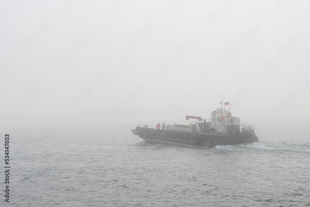 A barge transports a truck across a river. Poor visibility in fog. Passenger and automobile water transportation on a barge. Yana river, Magadan region, Siberia, Far East of Russia. Foggy weather.