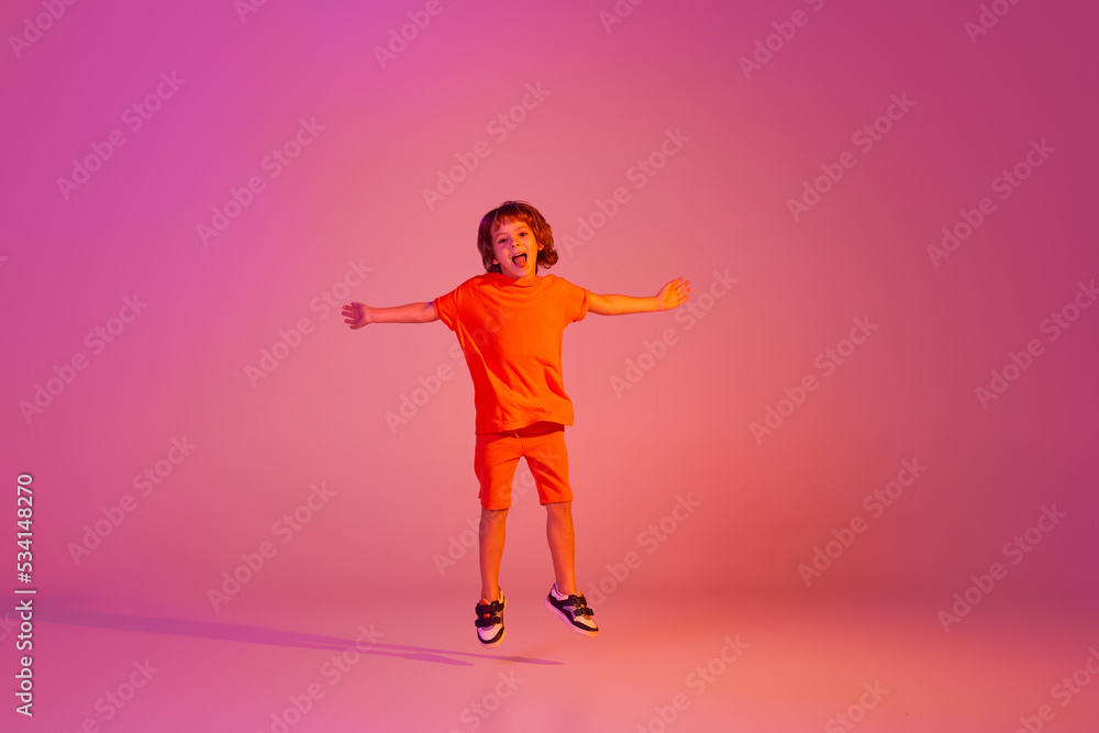 Portrait of caucasian little boy, kid in casual bright clothes dancing isolated over pink background in neon. Action, dance, happy childhood