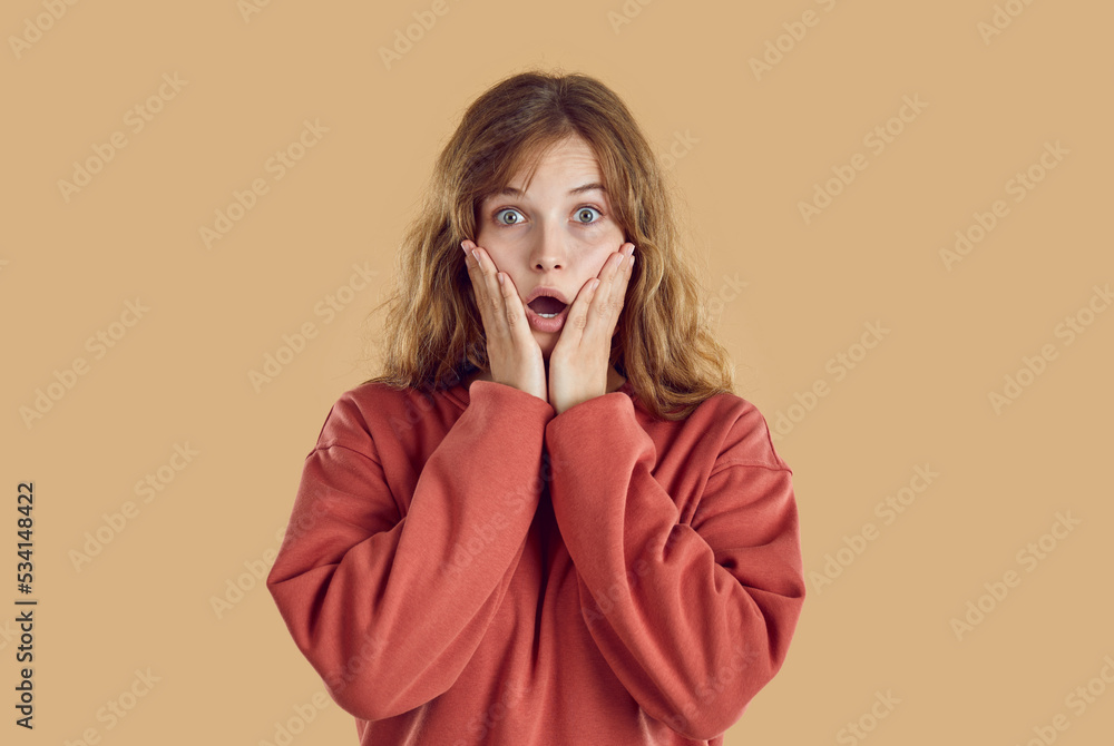 Portrait of young amazed woman with wide opened eyes and open mouth with hands on face. She is wearing red sweatshirt standing on beige background and looking at camera. Human emotions concept. Portrait of young amazed woman with wide opened eyes and open mouth with hands on face. She is wearing red sweatshirt standing on beige background and looking at camera. Human emotions concept.