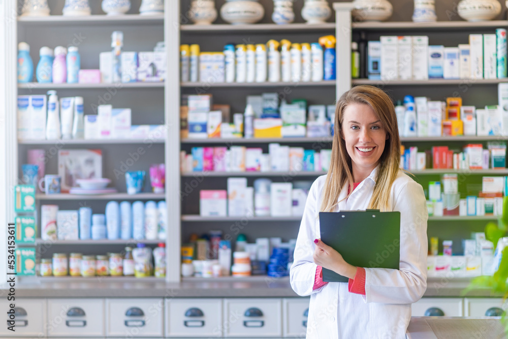 Young female pharmacist at work. Blurred shelves with pharmaceutical products on background. Pharmacist chemist woman standing in pharmacy - drugstore
