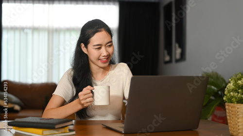 Pretty young woman drinking hot tea and working online with laptop computer on wooden table
