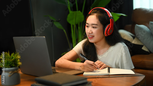 Concentrated woman in headphone listening lecture during study online on laptop and making notes on notebook