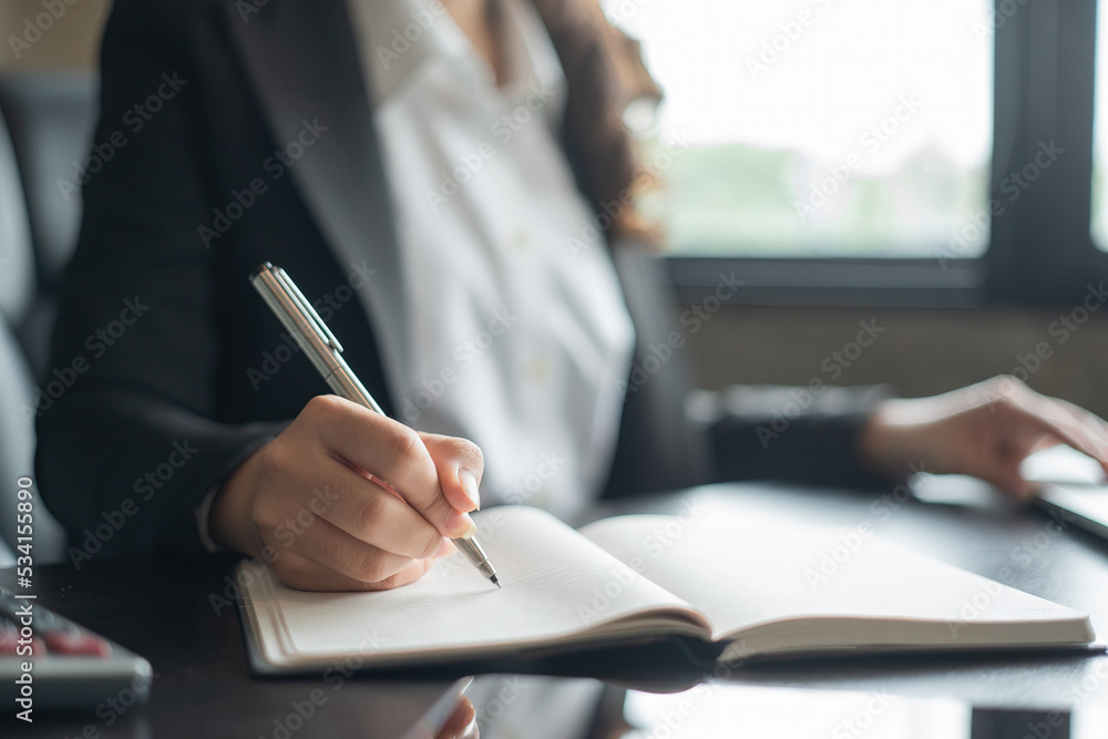 Close shot of businesswoman's hand holding a pen writing something on the paper in the foreground in office. Close shot of businesswoman's hand holding a pen writing something on the paper in the foreground in office.