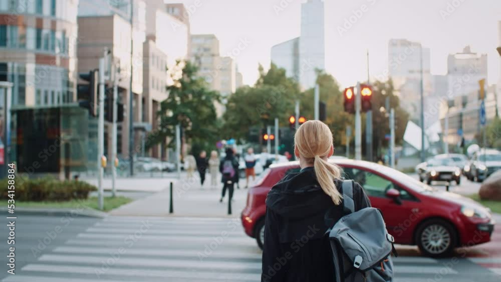 Young blond hair woman with backpack dressed in casual clothes standing by crossroad with lot of cars and pedestrians in city landscape. Female in busy street in evening, urban life, after work time Young blond hair woman with backpack dressed in casual clothes standing by crossroad with lot of cars and pedestrians in city landscape. Female in busy street in evening, urban life, after work time