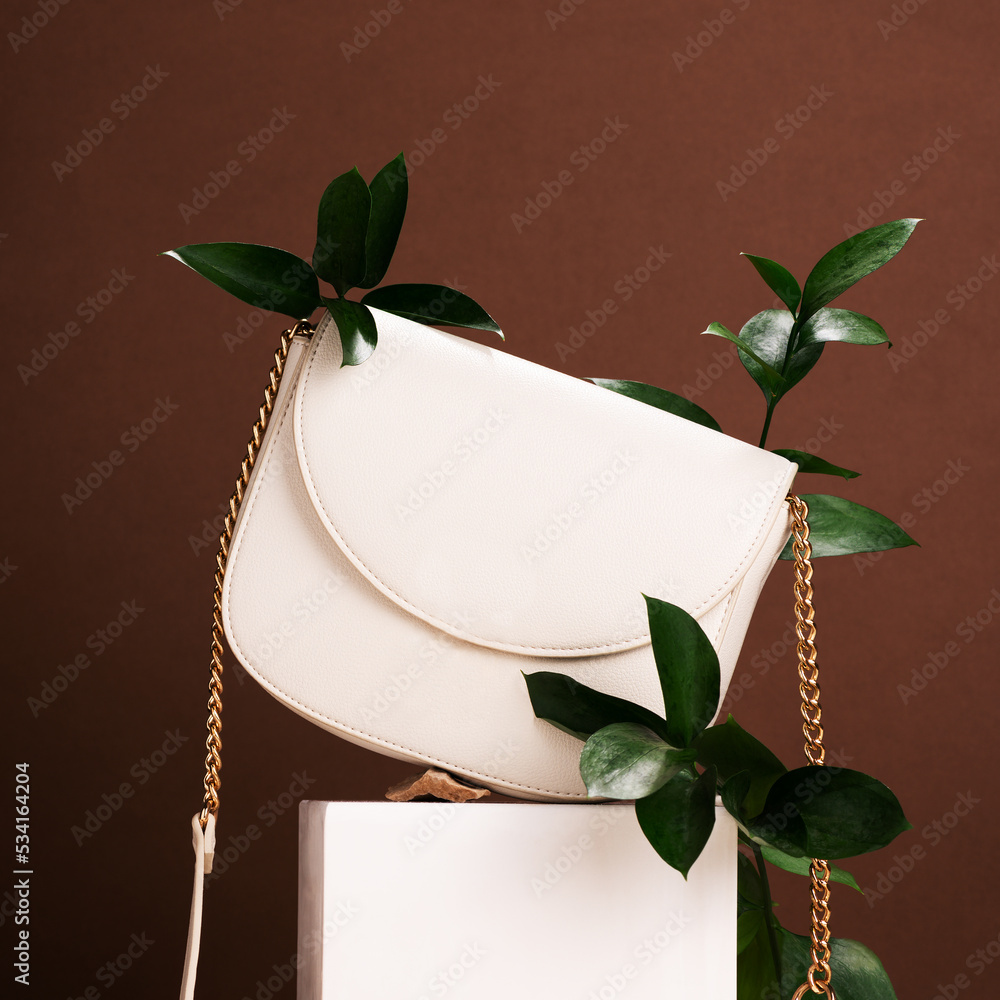 Stylish handbag with green branches on podium against brown background. Glamorous accessory for women presentation. White leather lady bag