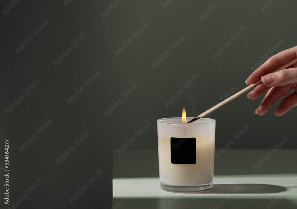 Woman lights candle in glass holder with wooden skewer on green background closeup. Candlelight creating cozy atmosphere at home. Copy space