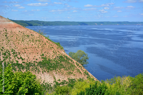 slope of mountain with red mud and green bushes and blue river on background