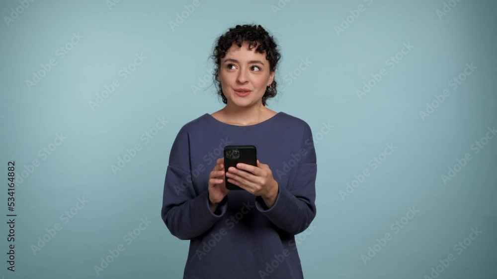 Smiling curly woman using smartphone typing browsing thinking about buying in an online store. Female holding mobile phone makes decision about an online purchase on isolated light blue background Smiling curly woman using smartphone typing browsing thinking about buying in an online store. Female holding mobile phone makes decision about an online purchase on isolated light blue background