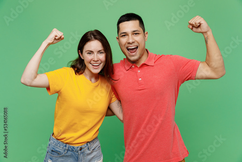 Young fun couple two friends family man woman wear basic t-shirts together doing winner gesture celebrate clenching fists say yes isolated on pastel plain light green color background studio portrait.