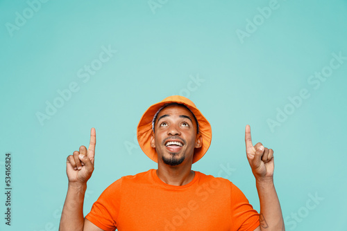 Young happy man of African American ethnicity 20s wear orange t-shirt hat point index finger overhead indicate on workspace area copy space mock up isolated on plain pastel light blue cyan background.