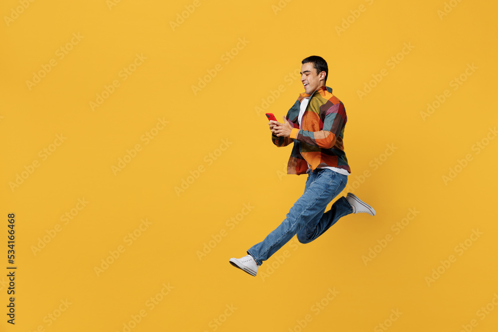 Full body side view fun young middle eastern man 20s wear casual shirt white t-shirt jump high run fast hurrying hold in hand use mobile cell phone isolated on plain yellow background studio portrait.