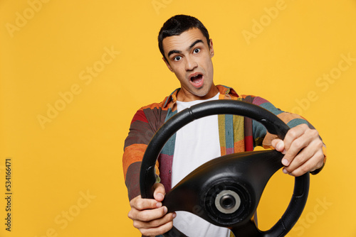 Young shocked fun middle eastern man he wear casual shirt white t-shirt pretending driving hold in hand steering wheel look aside on workspace area isolated on plain yellow background studio portrait.