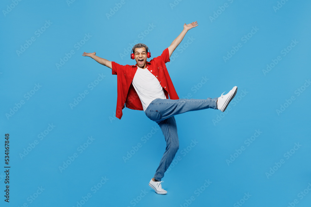 Full body young man of African American ethnicity 20s he wear red shirt headphones listen music dance raise up hands leg isolated on plain pastel light blue cyan background. People lifestyle concept.