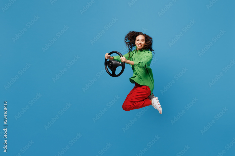 Full body side view young smiling fun woman of African American ethnicity 20s wear green shirt hold steering wheel pretending to drive car jump high isolated on plain blue background studio portrait.