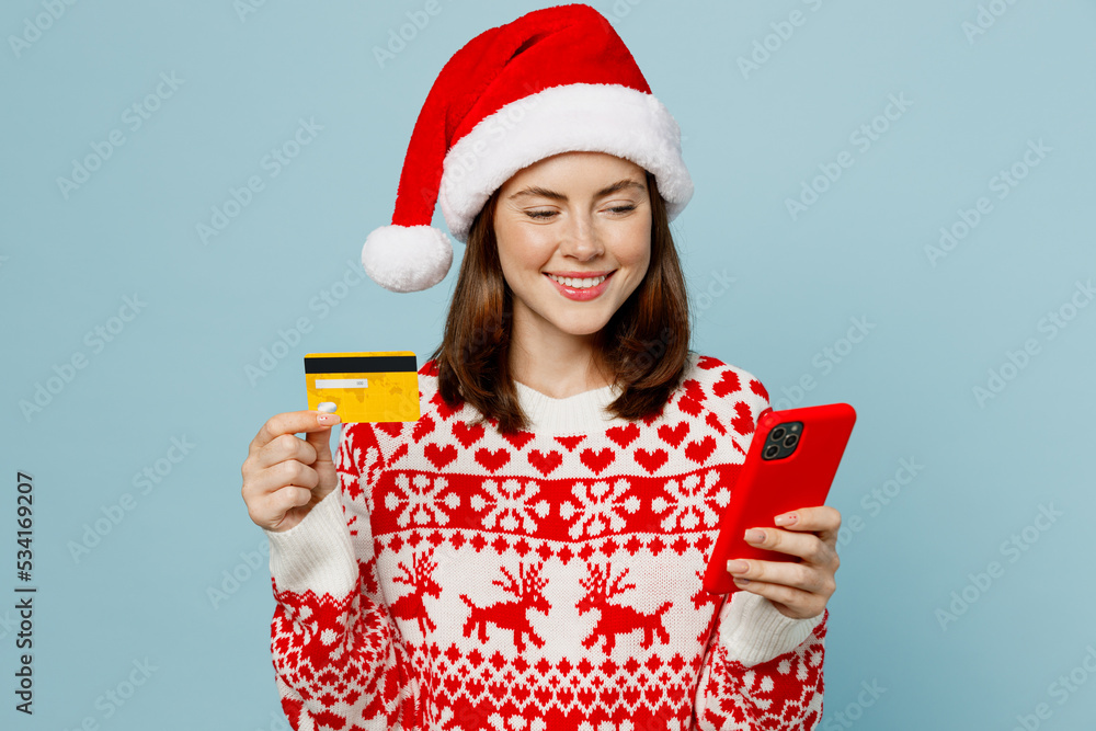 Young woman wear red knitted sweater Santa hat posing using mobile cell phone credit bank card order delivery isolated on plain pastel light blue cyan background. Happy New Year 2023 holiday concept. Young woman wear red knitted sweater Santa hat posing using mobile cell phone credit bank card order delivery isolated on plain pastel light blue cyan background. Happy New Year 2023 holiday concept.