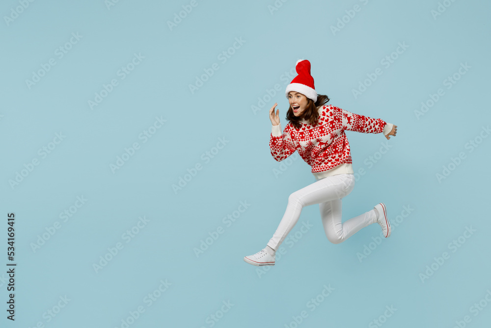 Full body fun young merry woman 20s wear warm red knitted sweater Santa hat posing jump high run fast hurry up isolated on plain pastel light blue cyan background. Happy New Year 2023 holiday concept.