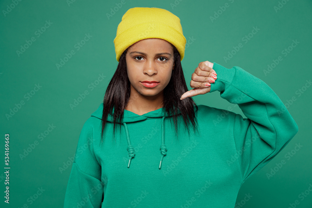 Little displeased sad kid teen girl of African American ethnicity 13-14 years old wears casual hoody hat showing thumb down dislike gesture isolated on plain dark green background Childhood concept.