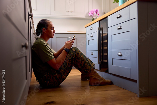Depressed Female Soldier Suffering With PTSD Sitting On Kitchen Floor On Home Leave Calling Helpline