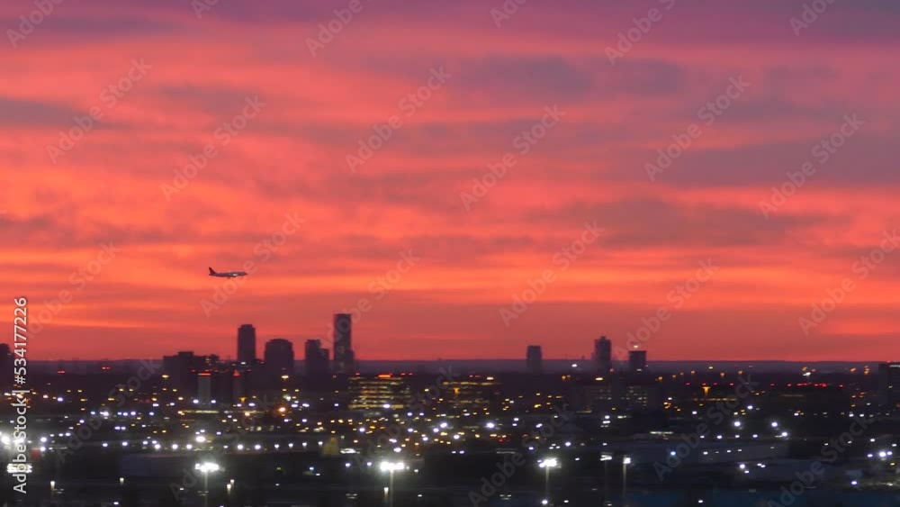 Silhouette of plane landing above dusk cityscape with glowing lights and red sunset sky Silhouette of plane landing above dusk cityscape with glowing lights and red sunset sky