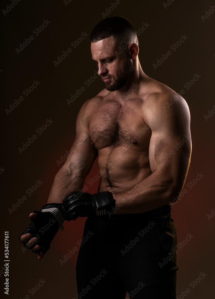 Studio portrait of fighting muscular man puts on fighting gloves posing on dark background. The concept of mixed martial arts. Brutal bodybuilder energy and power boxing.