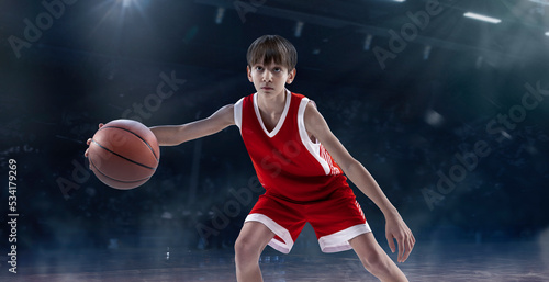 Portrait of teen boy, professional basketball player playing, dribbling isolated over sport stadium background