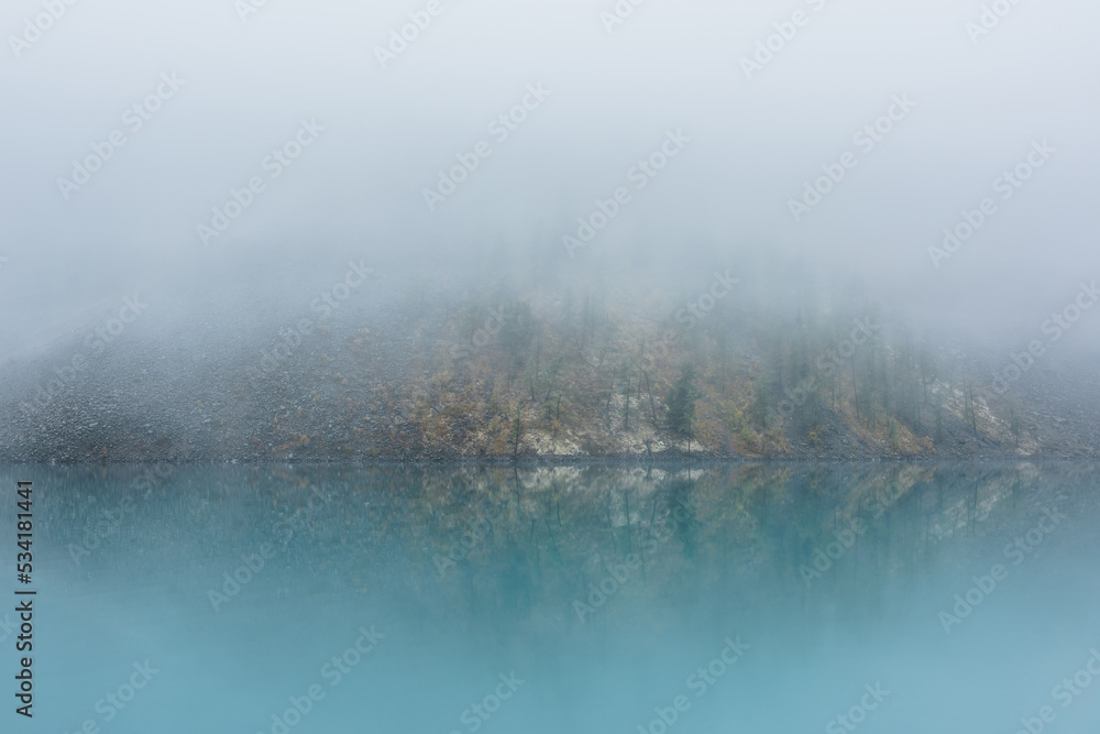 Fototapeta premium Simple meditative landscape with turquoise lake in thick fog. Tranquil view through clearance in dense fog to steep slope in fading autumn colors and mirror mountain lake. Reflective azure alpine lake