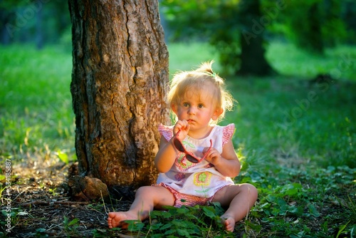 A baby girl is sitting in the forest on the grass and playing with sunglasses