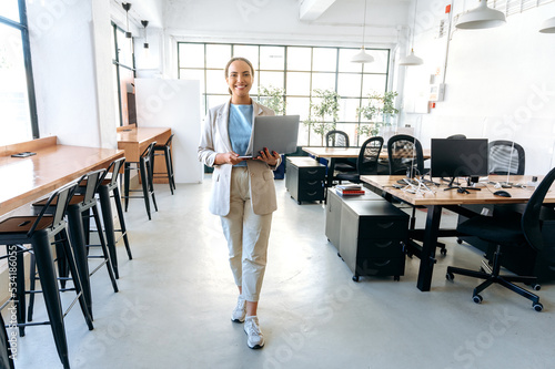 Modern trendy confident successful young mixed race woman, wearing stylish business clothes, go across the creative office, holding an open laptop in her hands, looking at the camera, smiling