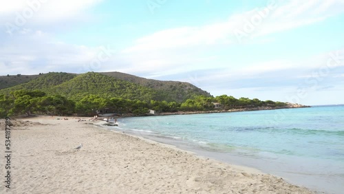 The waves splashing on beach coastline of Cala Agulla (Cala Ratjada) on Mallorca in the mediterranean sea. Deep blue water and a green pine forest in the background. People bathing,  4k.