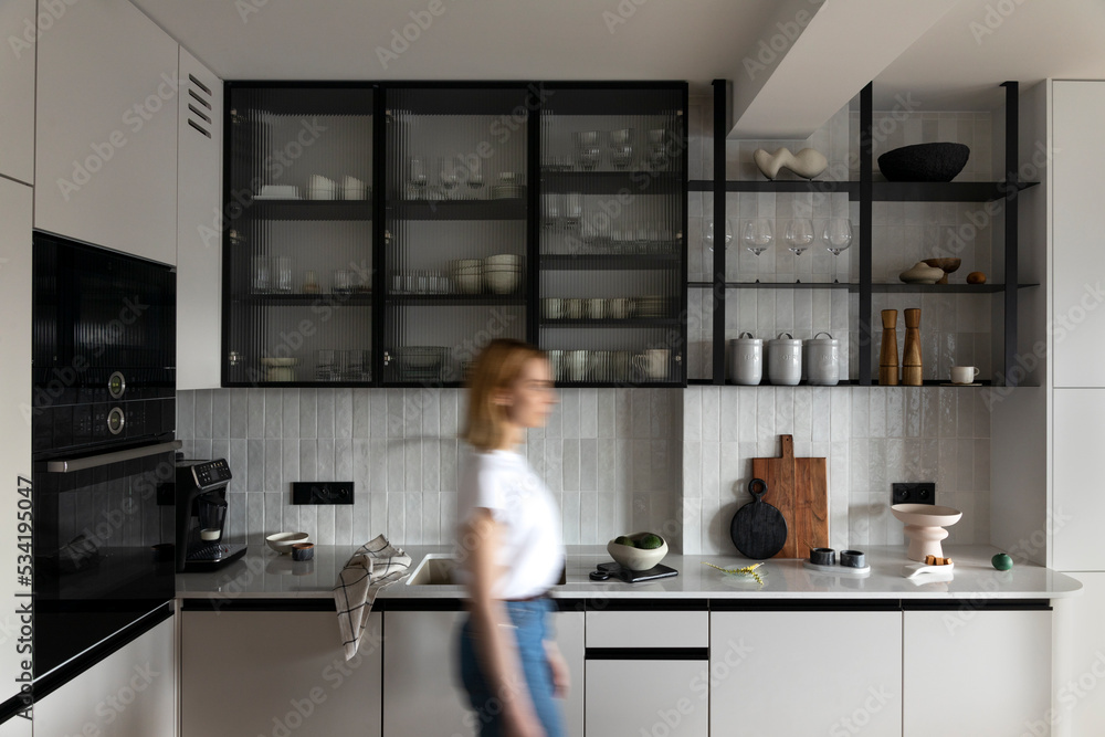 Modern composition of dining space with design kitchen island, furnitures, kitchen stuff, cup of coffee and elegant personal accessories. Woman walking through space. Stylish home decor. Template.  