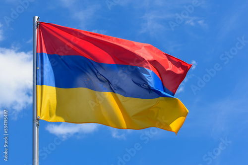 The flag of Armenia is a rectangular banner consisting of horizontal red, blue and orange stripes on a background of blue sky during the summer day. photo