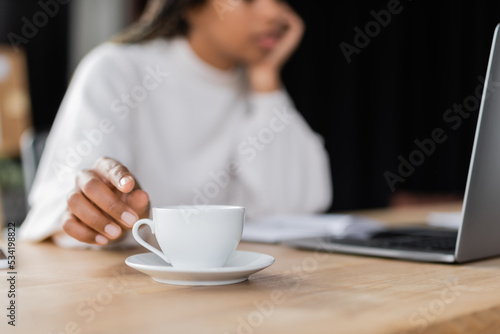 Cropped view of african american businesswoman taking cup of coffee near laptop on table.
