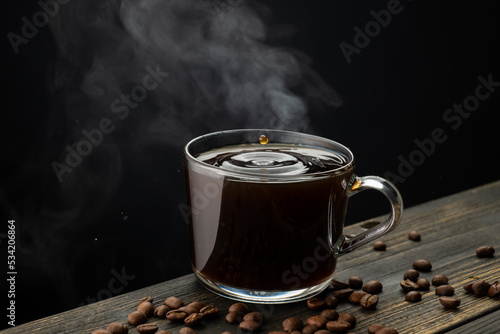 A mug of hot coffee with evaporation and steam and a small splash on a wooden table next to coffee beans