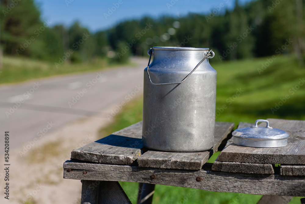 Aluminum vintage milk can by the road in summer. The lid lies on a wooden bridge. Collection of milk from farmers in the countryside. Latvia